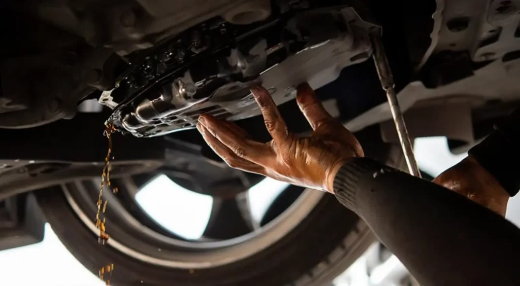 A person's hand reaching under a vehicle, possibly pointing to a transmission component or a leak, with the undercarriage visible