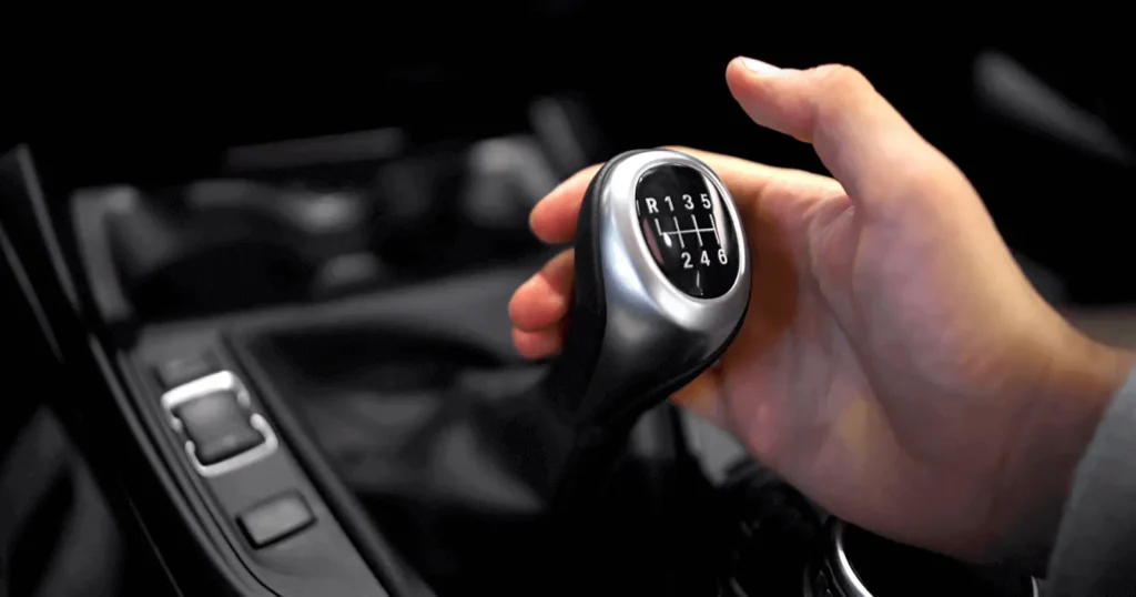 A close-up of a hand gripping a manual gear shifter, displaying the "R 1 3 5" and "2 4 6" pattern.