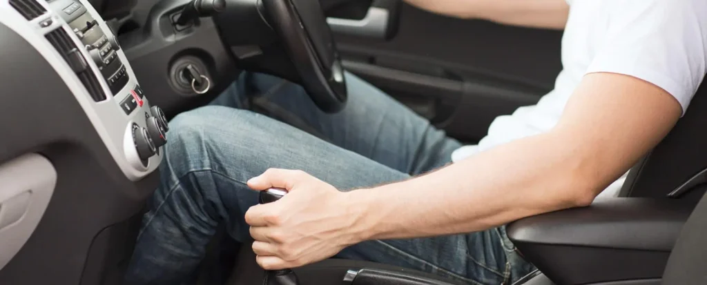 A person's legs and right arm inside a car, with their right hand on the manual gear shifter and their left foot near the clutch pedal, demonstrating proper driving posture.